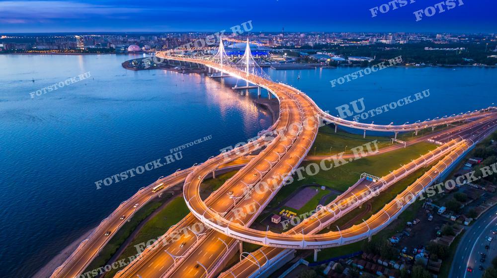 На фотографии изображен вантовый мост через реку Неву в Санкт-Петербурге в сумеречное или раннее утреннее время. Мост освещен, и его огни создают динамичное ощущение движения, в то время как свет отражается в воде, добавляя красоты городскому пейзажу. Это изображение подходит для демонстрации современной городской инфраструктуры и может быть использовано в информационных брошюрах, образовательных материалах, а также как элемент дизайна интерьеров.